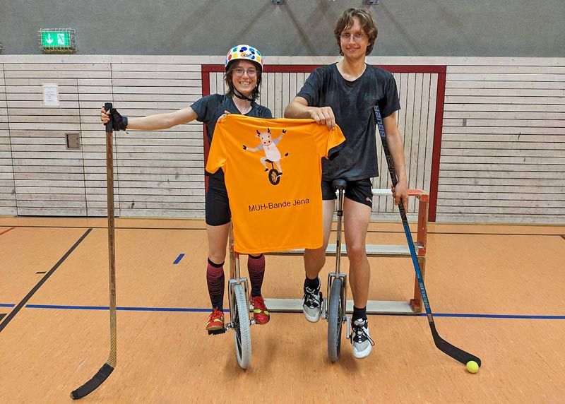 Lisa Hils (l.) und Fin Thiessen posieren in der Einradhockeytrainingshalle vor einem improvisierten Hockeytor und halten stolz ihr erstes Team-Trikot in die Kamera.