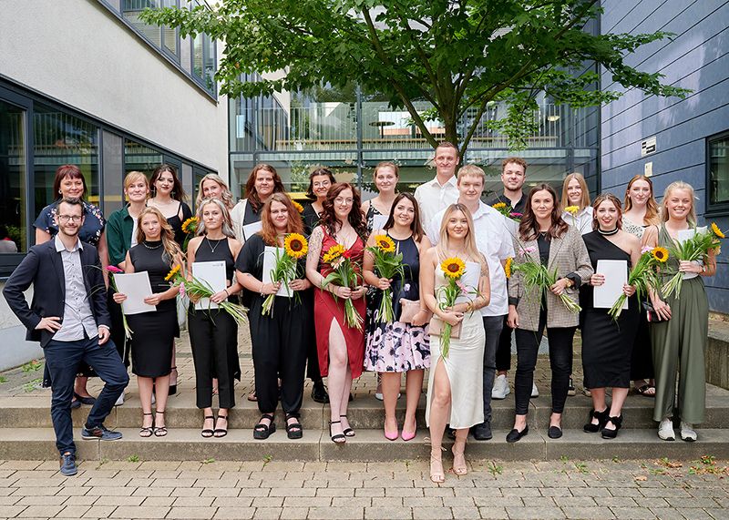 Freudestrahlend halten die Absolventinnen und Absolventen der generalistischen Pflegeausbildung am Universitätsklinikum Jena Zeugnisse und Sonnenblumen in den Händen.